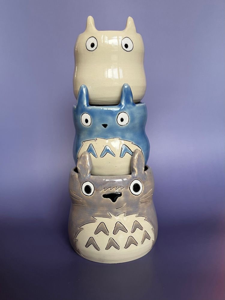 Ceramic cup set featuring characters from the movie “My Neighbor Totoro”. The cups are stacked, with the bottom and largest cup being the character Totoro. Totoro is grey with a white belly that has grey markings inside. Totoro is smiling with whiskers, a black nose and tall straight ears that extend up above its eyes. The middle cup is based on the character “blue totoro”. This character cup is blue with a white belly, a small triangle nose and ears that point straight up. At the top is the third character cup, “white totoro”, who is colored white, only has eyes and small pointy ears that point upwards. These cups are stacked on top of each other, and get smaller as they progress up. 