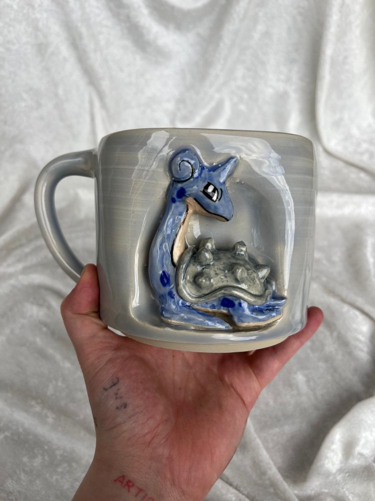 Lapras pokemon sculpture ceramic mug. Lapras is on the outside center of the mug, and the mug is colored an icy light blue
