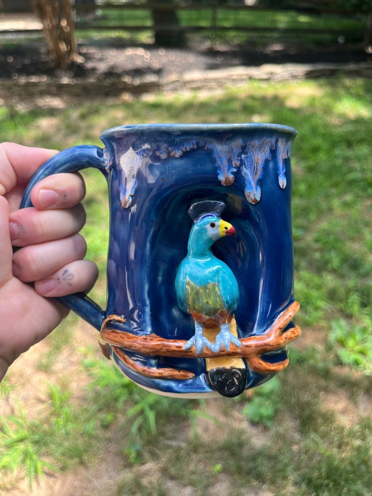 Great blue turaco (bird) ceramic sculpture mug. The bird is perched on a branch on the outside center of the mug. The mug is colored a dark blue with white glaze drips on the outside rim. The bird is primarily a bright blue color with a green stomach, a yellow and red beak, and a dark blue array of head feathers