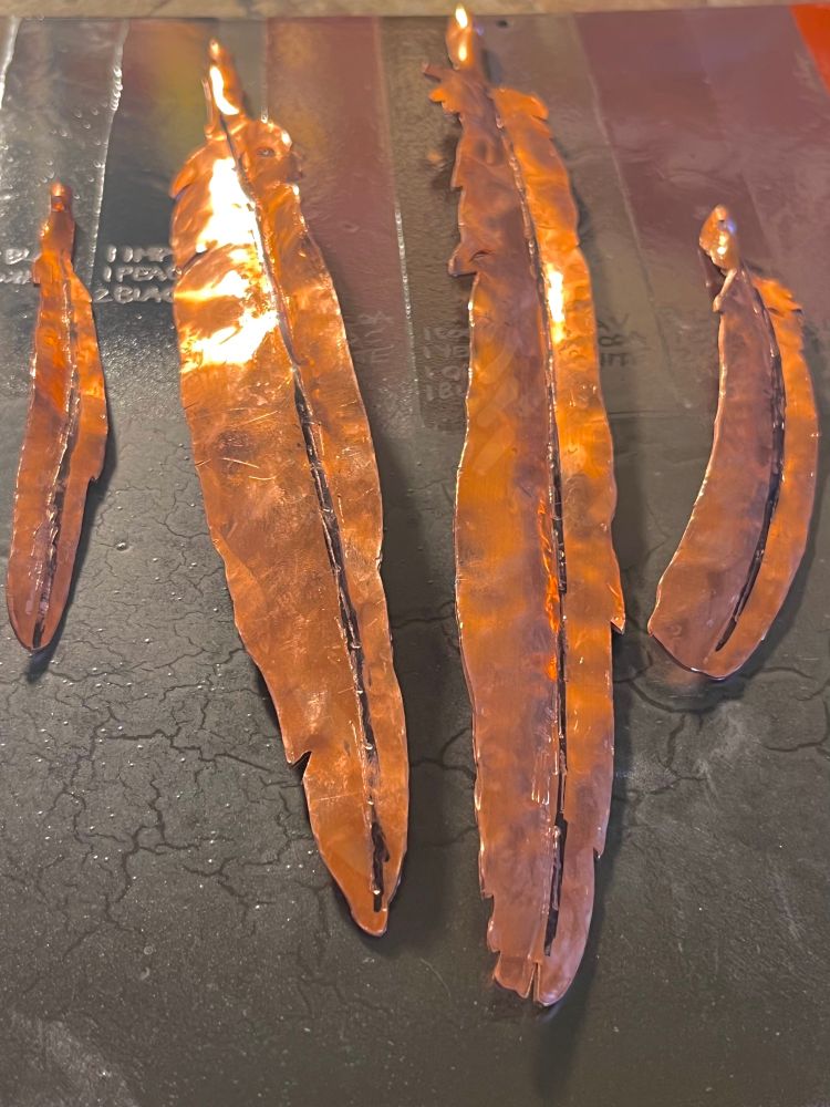 Four different feathers made of hammered copper await enamel colors. Once completed, the matching feathers will be attached with wire to the enameled panels with images of a Belted Kingfisher, a Black-billed Magpie, an American Crow, and a Western Bluebird.