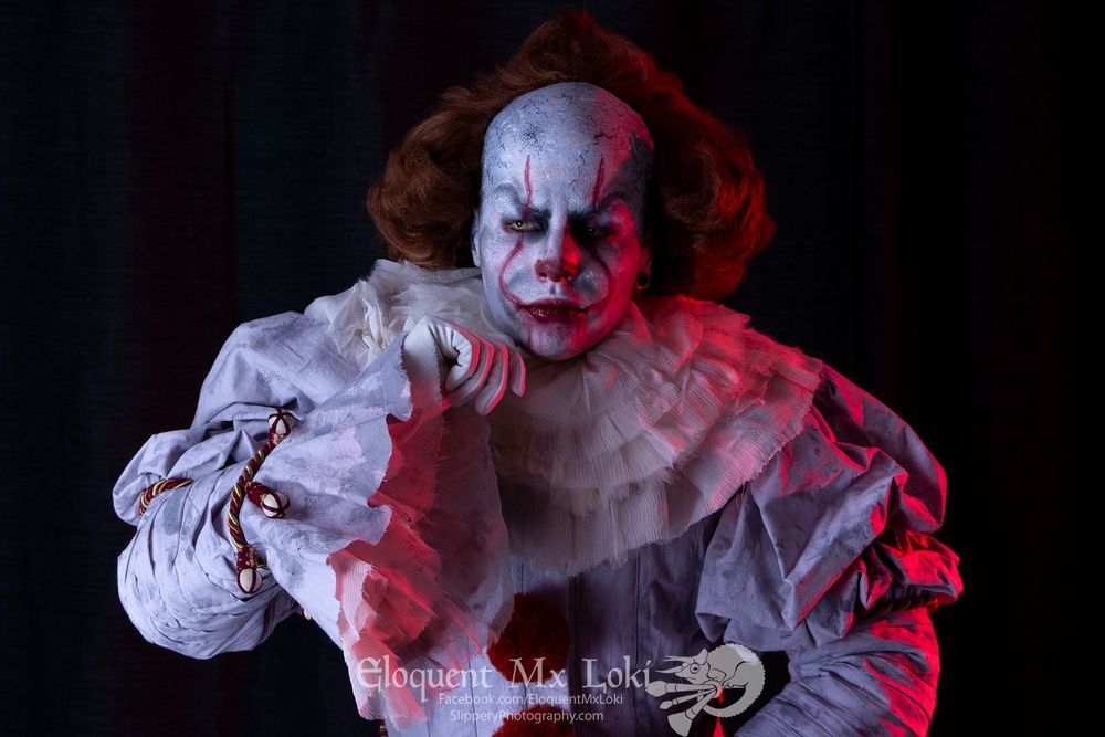 Mx Loki is cosplaying as Pennywise the dancing clown from the horror film It inspired by the novel by Stephen King. There is a spooky red light flashed across one side of the photo and Loki has one hand posed near their chin.