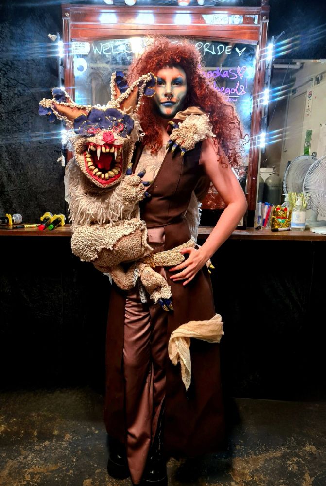 Loki has on a curly ginger wig and bold blue makeup. They have a monster (puppet) called Tiebold sitting on their hip with his arm around their shoulders. Tiebold has a big mouth full of teeth, two sets of ears, a shaggy body and a bat-like nose. They are standing infront of a beauty mirror with lights casting a halo of little rainbows around them.