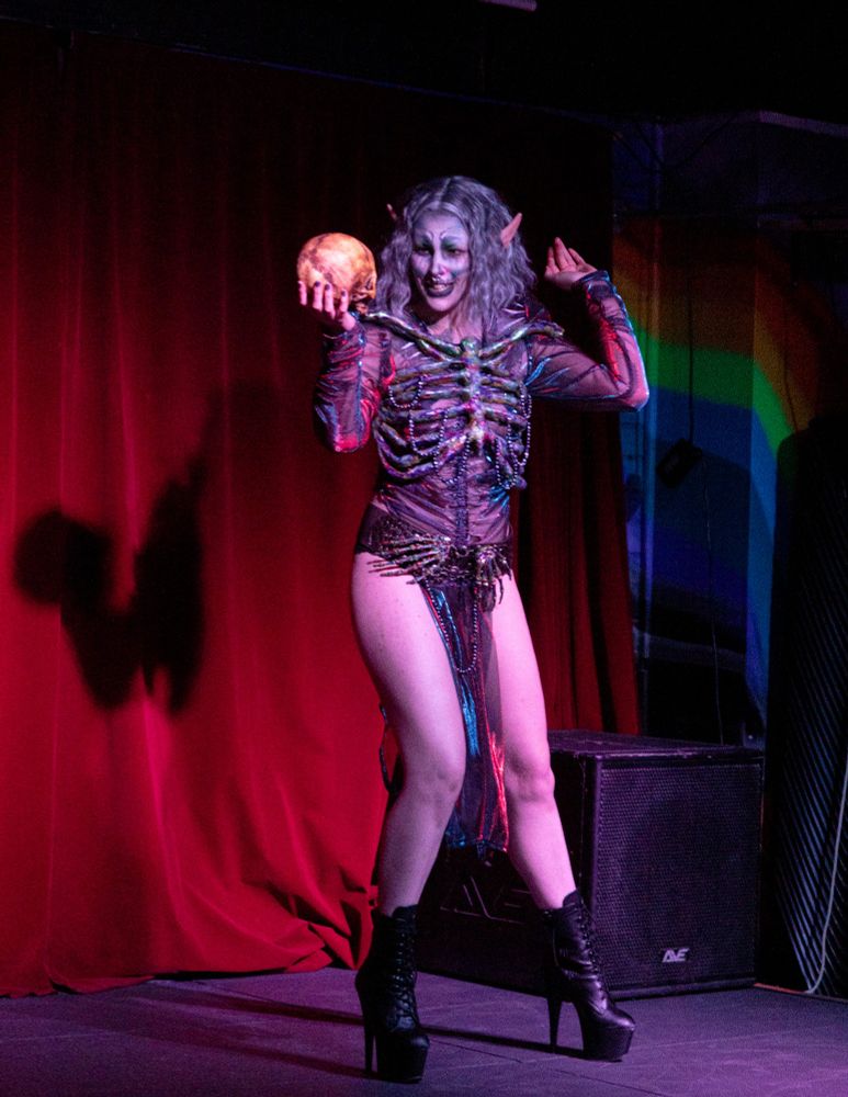 Loki is on stage dressed as an Elf Necromancer, wearing a silver wig and skull inspired makeup. They have on a see through duochrome tunic over which is an opalescent ribcage chest piece and a belt with opalescent bone hands. They are smiling at the skull the are holding.