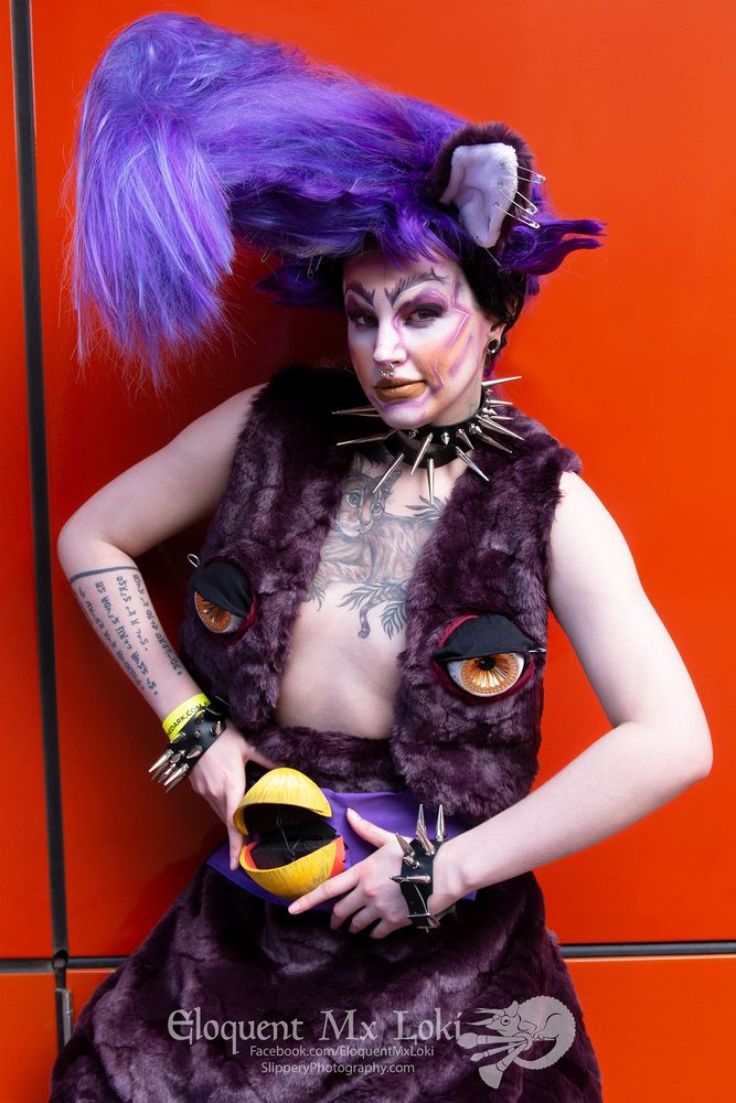 Loki is in a purple fur vest with furby eyes on the chest and pants with a furby beak. They have a big purple pompadour wig with purple furby ears in it, the ears have safety pins as piercings. Loki is also wearing large spikey leather wrist bands and choker.