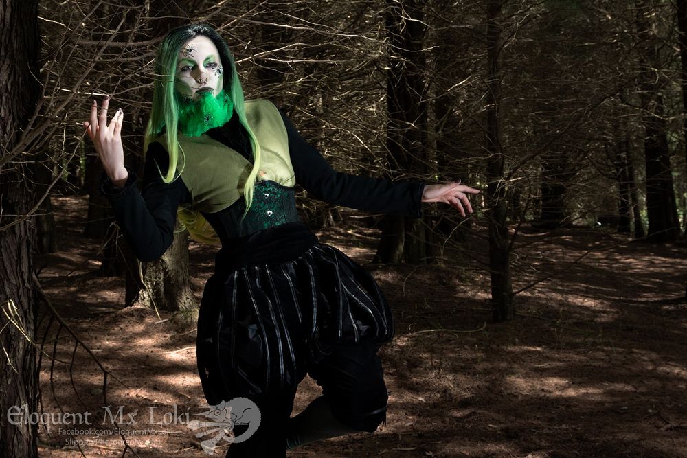 Mx Loki is modeling black puff pants a green vest under a green corset and is wearing a long gradient green wig and a green beard. They have pale makeup with green accents and are posing in a forest.