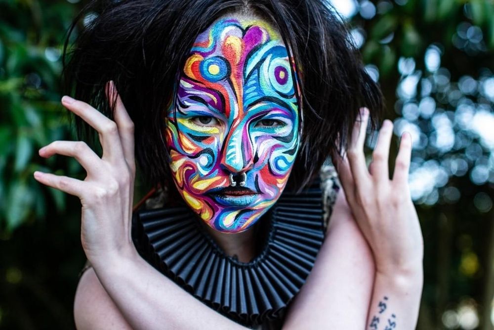 Mx Loki is staring into the camera, they have framed their face with their hands and are wearing a leather clown neck ruffle. Their face is painted by artist Jessie Ngaio with a magical swirl of vibrant colours that coalesce and form an abstract mask.