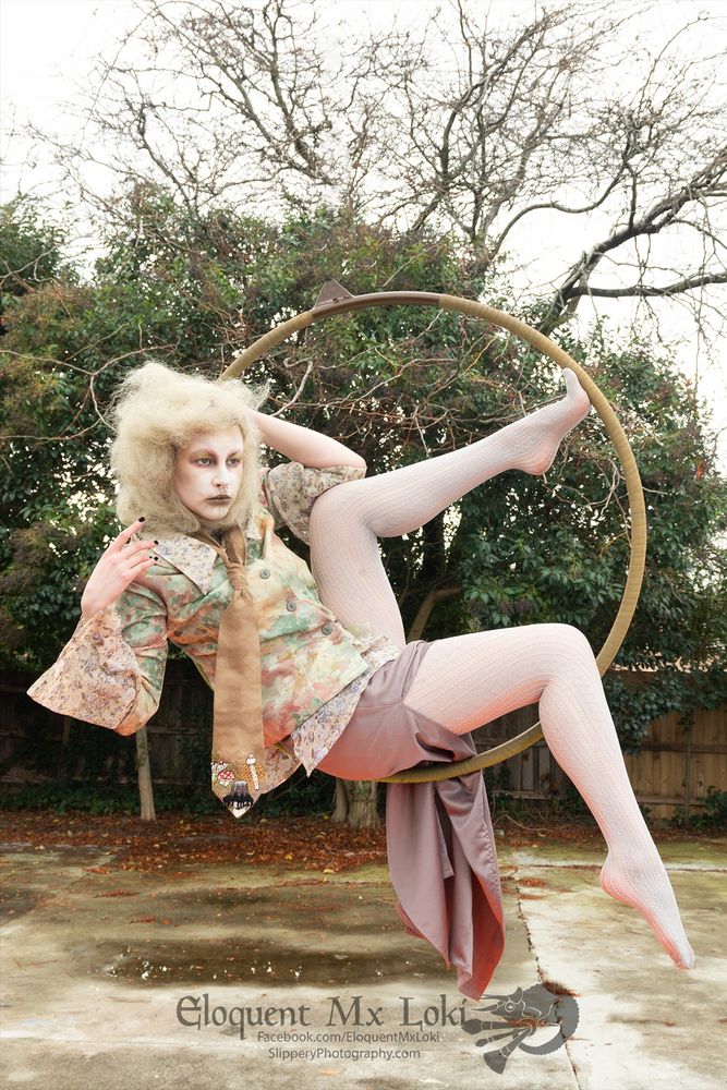 Loki is posing in an aerial hoop in a romantic floral ensemble. They have a pale blinde wig styled in a dreamy cloud style and desaturated makeup like an old faded painting. There are trees in the background.