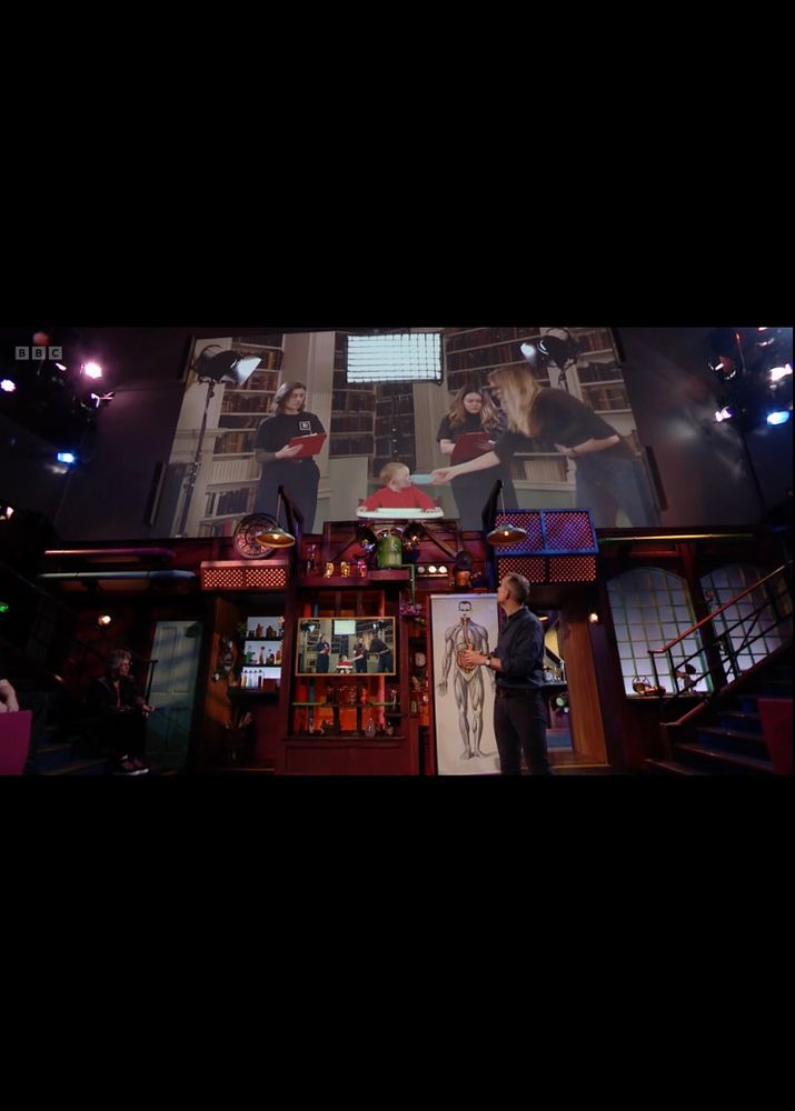 In the Royal Institution lecture theatre, Chris looks up at the projector feed where two adults, including me, are dressed as scientists with clipboards. Between us sits Chris' baby, Indigo, in a high chair, curiously trying unusual food.