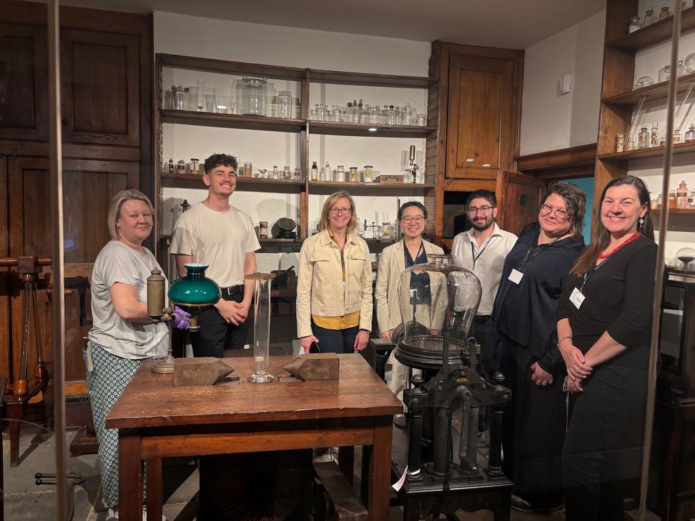 7 people behind the glass wall of Faraday’s original laboratory, surrounded by his apparatus 