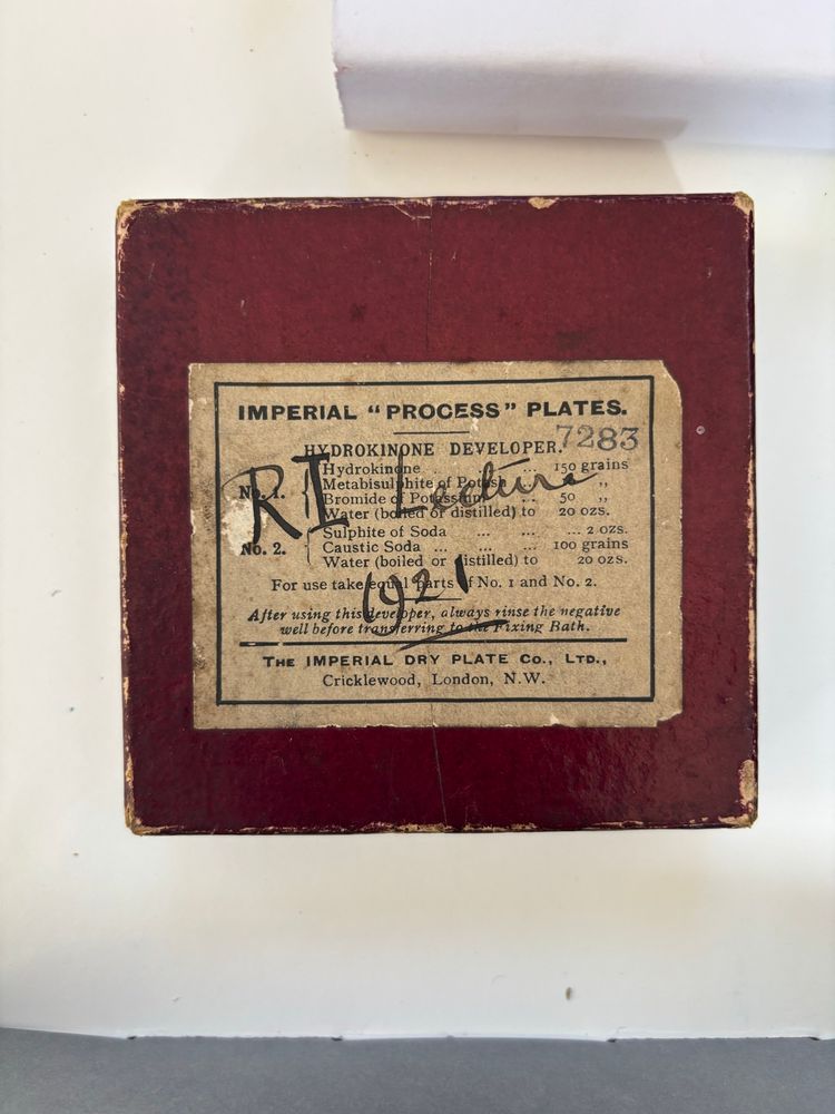 Top down view of a red glass slide box, with “R.I Lecture 1921” written in ink over the yellowing label on top (containing information about the plate, Imperial “process” plates). The box is slightly worn around the edges. 