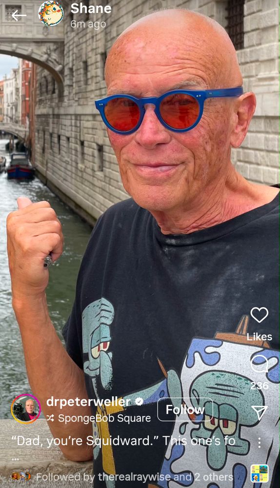 Peter Wellers wearing a tshirt, in what looks like Venice, a tshirt depicting Squidward painting Squidward.  Peter smirks at the camera and his thumb points back at the Venice canal 
