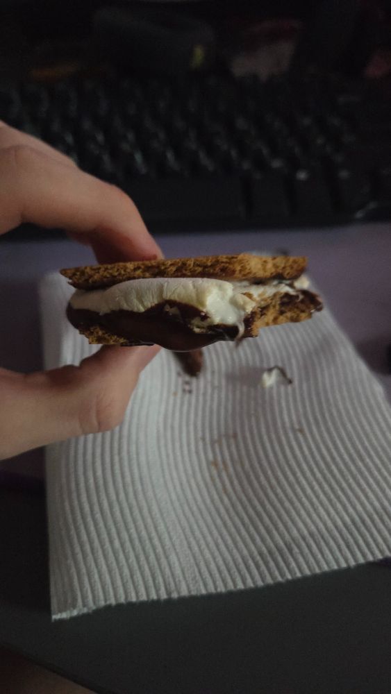 A s'more being held over a paper towel in front of a computer keyboard. There is some melted chocolate on the paper towel.
