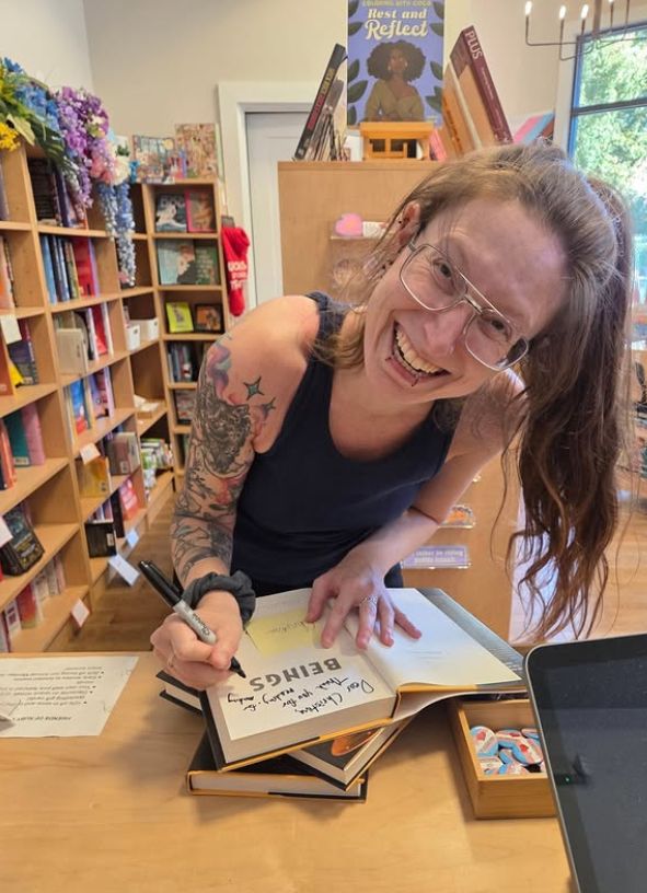 Me, a white queer with glasses and piercings and a tattoo sleeve, wearing a tank top, with my hair up in a high ponytail, smiling at the camera widely while signing a copy of BEINGS in All She Wrote Books.