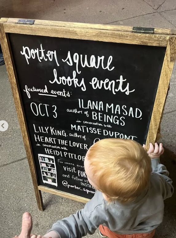 A redheaded toddler holding onto the chalkboard sign outside of Porter Square Books that includes the info about my event there