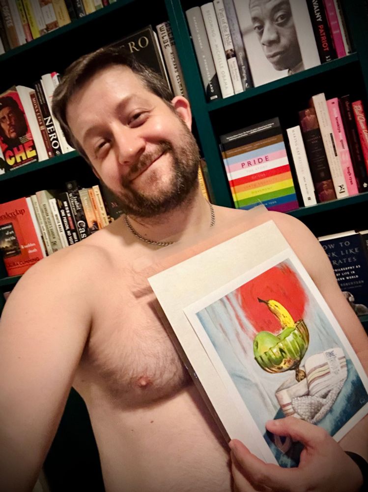 Naked bearded gay boy holds a print of his painting, “Supported Froot”, which features a BIKE jockstrap around an amber glass bowl with apples and bananas in it. A bookshelf is in the background. 