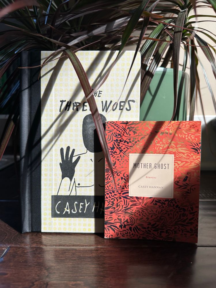 Photo of two books by Casey Hannan - “The Three Woes” and “Mother Ghost” - in front of a green houseplant. 