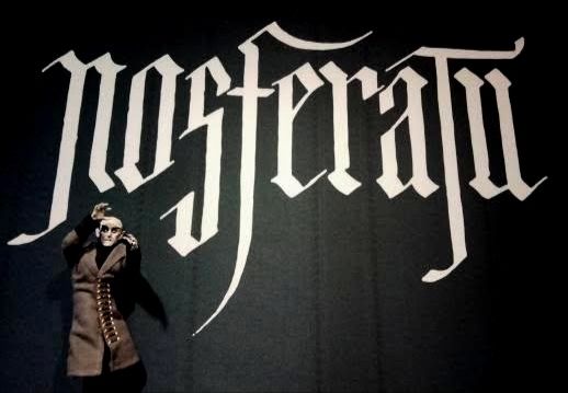 A toy of Count Orlok with raised arms, underneath the Nosferatu logo on a movie poster 