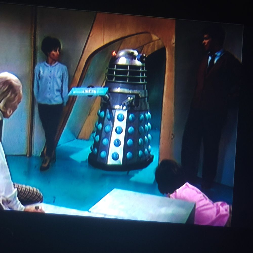A Dalek enters the room where the Doctor and companions are being held, holding out a blue tray of food.