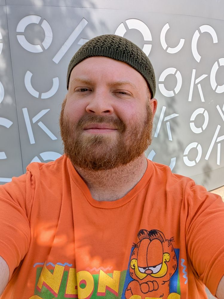 Very handsome red bearded man wearing a dark green knit cap and an orange shirt that says "Neon Genesis Evangelion" accompanied with a picture of Garfield the cat in front of an art installation featuring the letters "O, K, and C" in various positions across a metal surface which could be misconstrued to be seen as other words. 