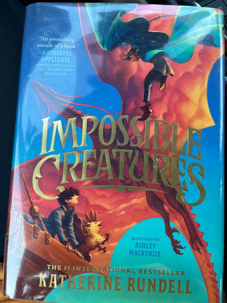 The cover for the book Impossible creatures by Katherine Rundell, showing a boy in at the front of a boat with a golden baby griffin, looking up at a red dragon and a flying girl.