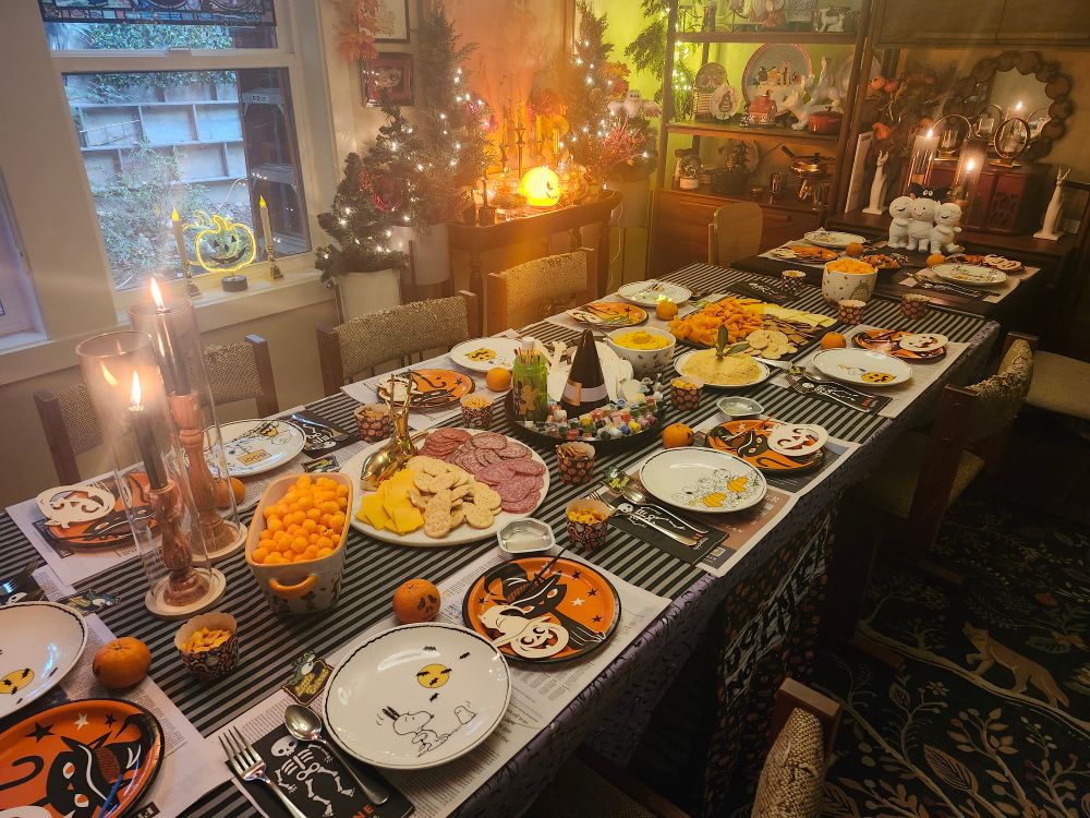 A dining room table with multiple settings with newspaper under a ceramic plate and a paper plate with a wooden craft to be painted, with crackers and cheese and carrots and other snacks in the middle, and a plastic witch's hat with paint pots around the brim