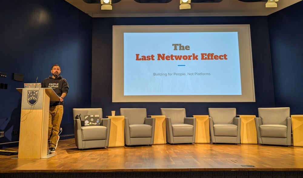 Anuj stands at a podium on a stage, behind him a screen displays the slide "The Last Network Effect"