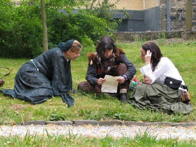 Farbfoto dreier Frauen, die in LARP-Kostümen auf einer Wiese sitzen und ein Dokument studieren.