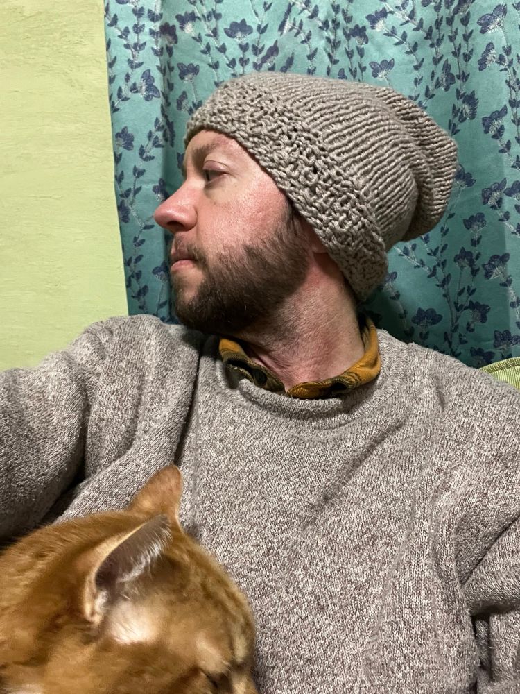 Photo of me on the couch with a gray-cream colored knitted hat on my head and my or age cat Kit on my lap (ears in the bottom left corner).