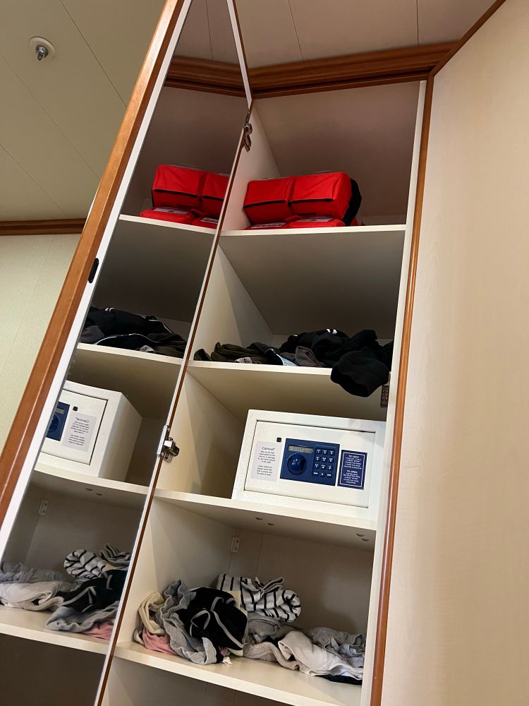A picture of a long narrow, cupboard with several large shelves, a cupboard door on the left with a mirror on it, and on the top shel, far above the reach of a wheelchair user, two lifejackets