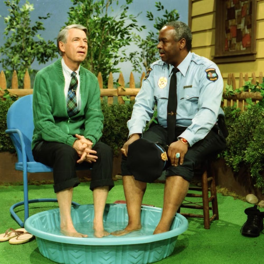 Mister Rogers and Officer Clemmons sit together, both of their feet are in the same tiny tub of water 