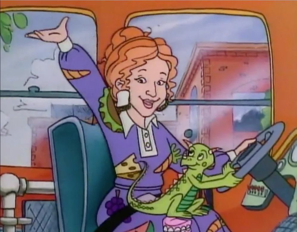 A smiling Ms. Frizzle, in colorful dress, sits in driver’s seat of The Magic School Bus. Liz, a smiling green lizard, sits in her lap holding the steering wheel.
