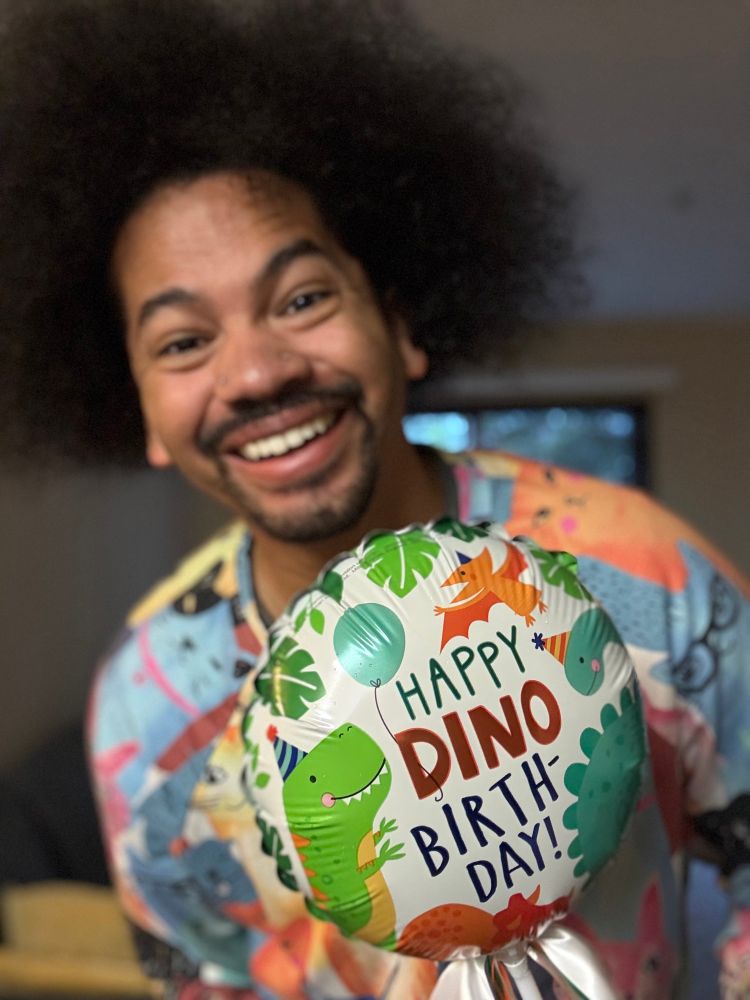 A blurry Mychal smiles with a focused shot on his birthday balloon reading, “Happy Dino Birthday-Day!” (with cartoon dinosaurs on the balloon)