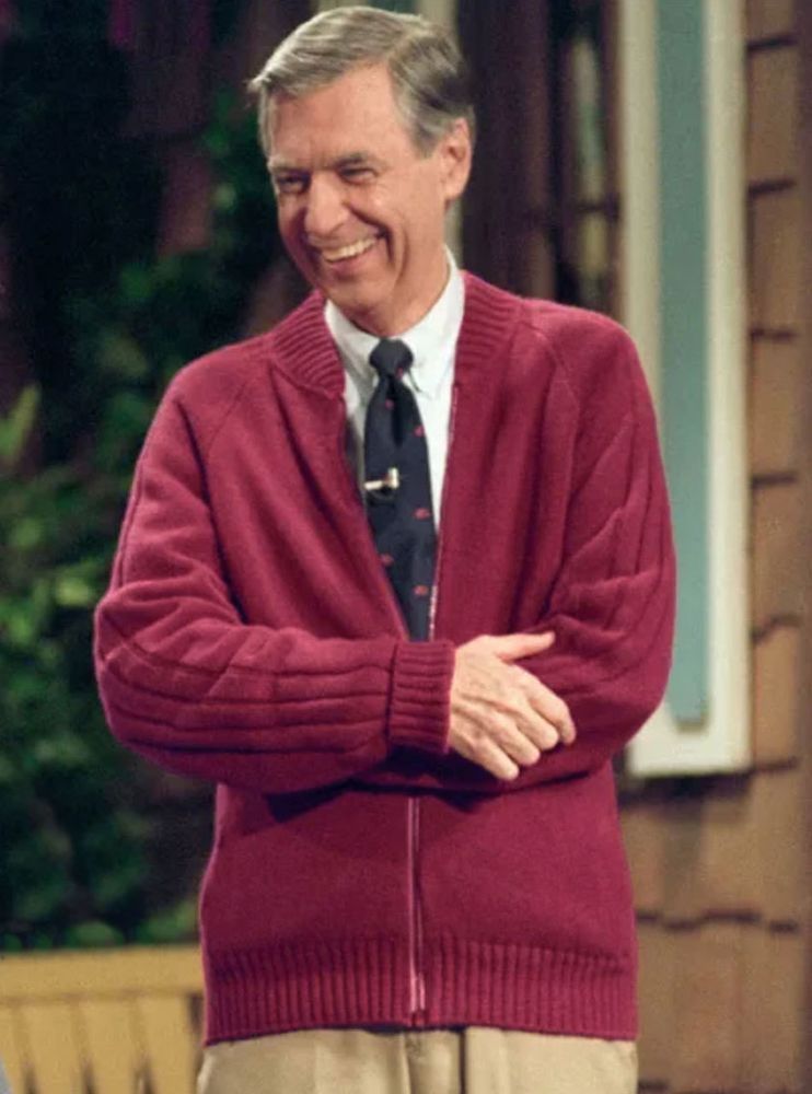 A smiling Mister Rogers crosses his arms. He wears a red cardigan and a black and red tie.
