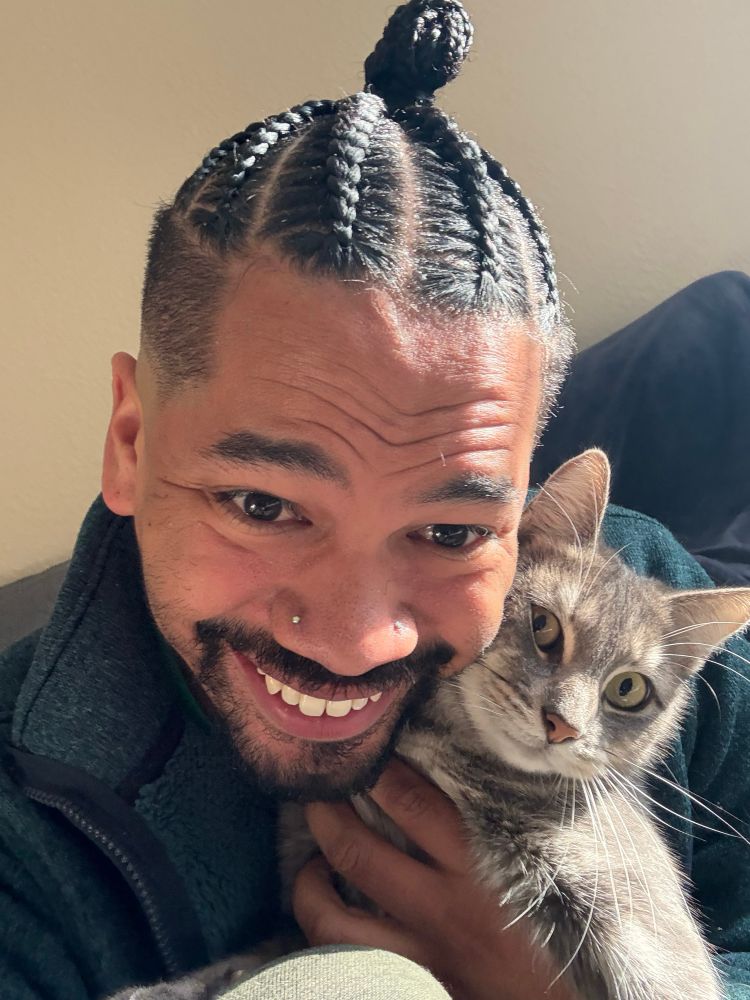 A smiling Mychal, with braided hair and a green jacket, holds his yellow-eyed gray cat (Machine Gun Kitty) next to his face