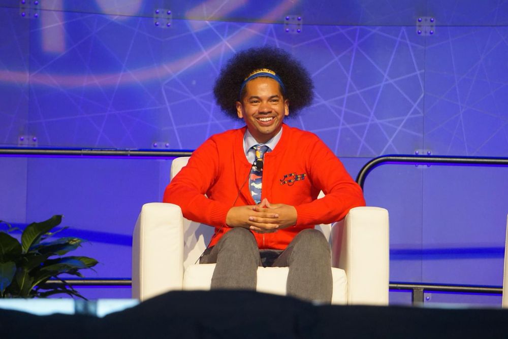 Mychal smiling, while seated and hands together. He wears a red cardigan and dinosaur tie.