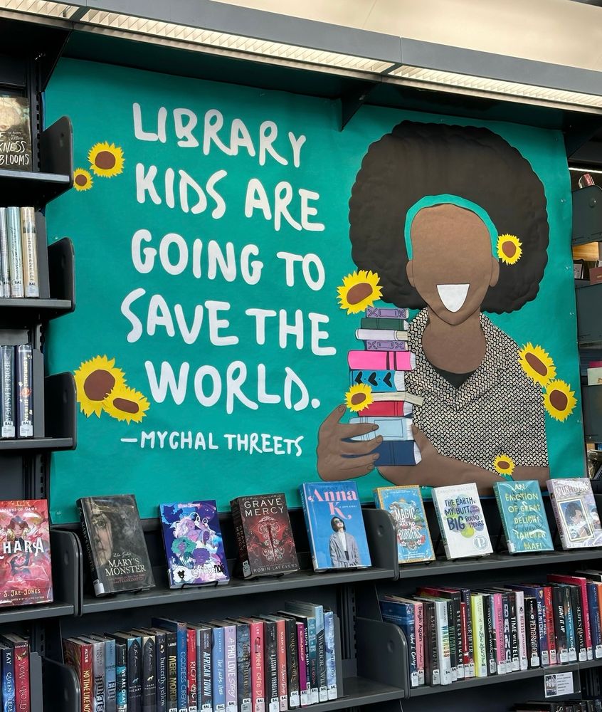 Library display based on artwork from Dev the Pineapple. A smiling Mychal, illustrated, with books and sunflowers. Text quote reads, 