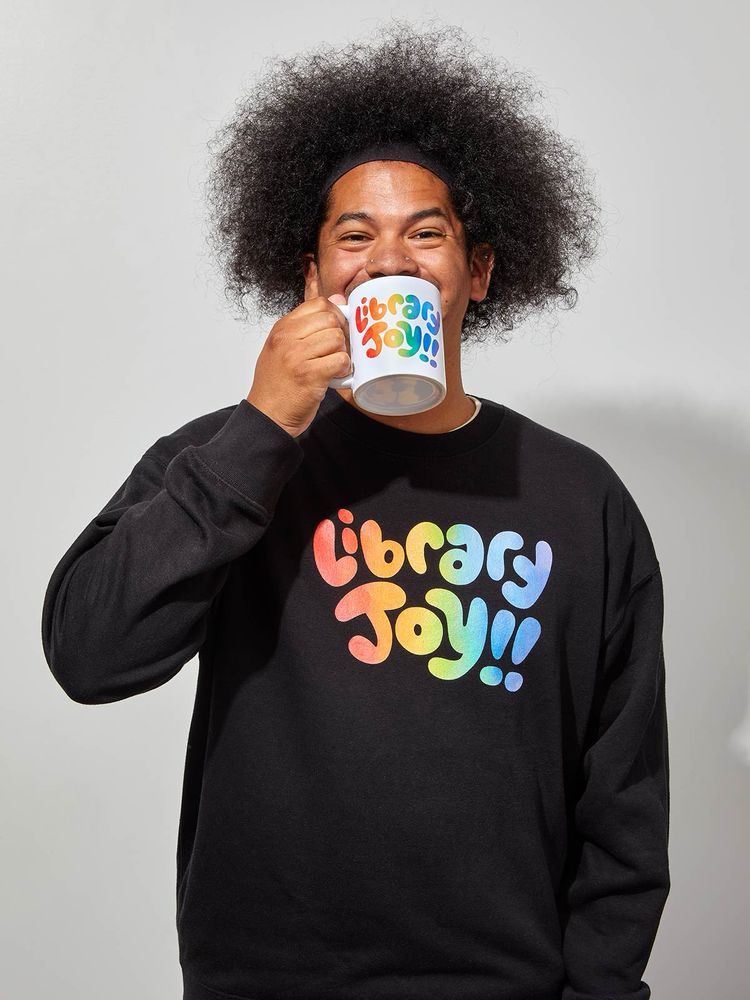 Mychal smiles while sipping from a mug that in colorful lettering reads, “Library Joy!!” His sweater reads the same message 