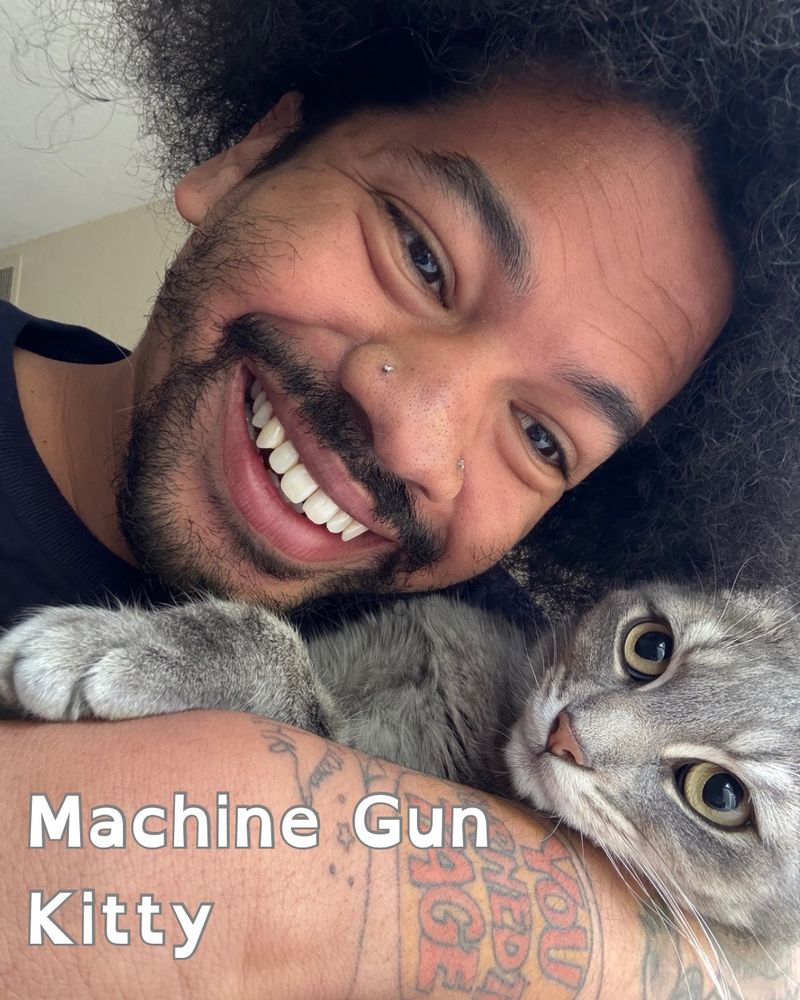 Mychal smiles and holds his yellow-eyed cat, who is grey and white, and goes by the name Machine Gun Kitty