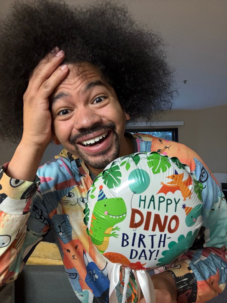 A shocked Mychal puts his hand on his head, holding his birthday balloon with animated dinosaurs that reads, “Happy Dino Birth-Day!”