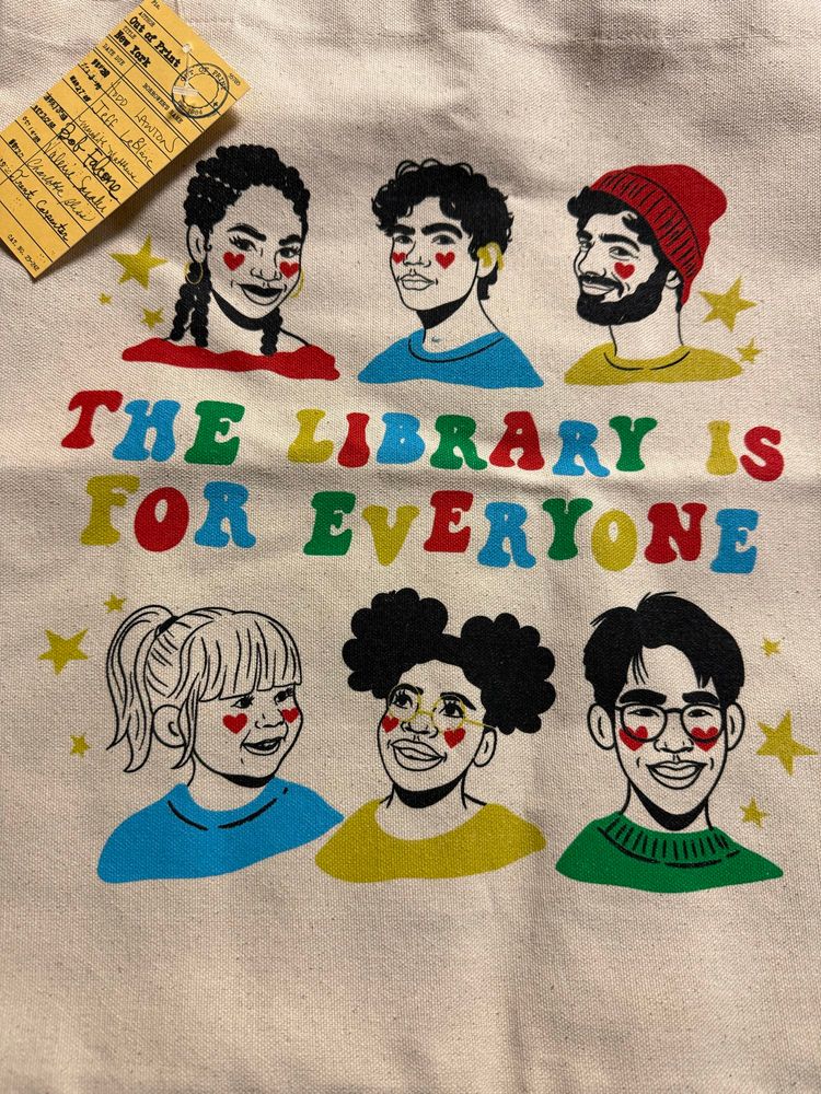 The Library is for Everyone tote bag from Out of Print, artwork by Dev the Pineapple. Art is of various faces, representing the library being for everyone.