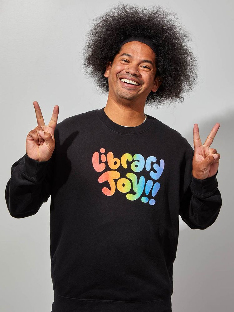 Mychal smiles while holding up two peace signs with his hands. His sweater in colorful lettering reads, “Library Joy!!”