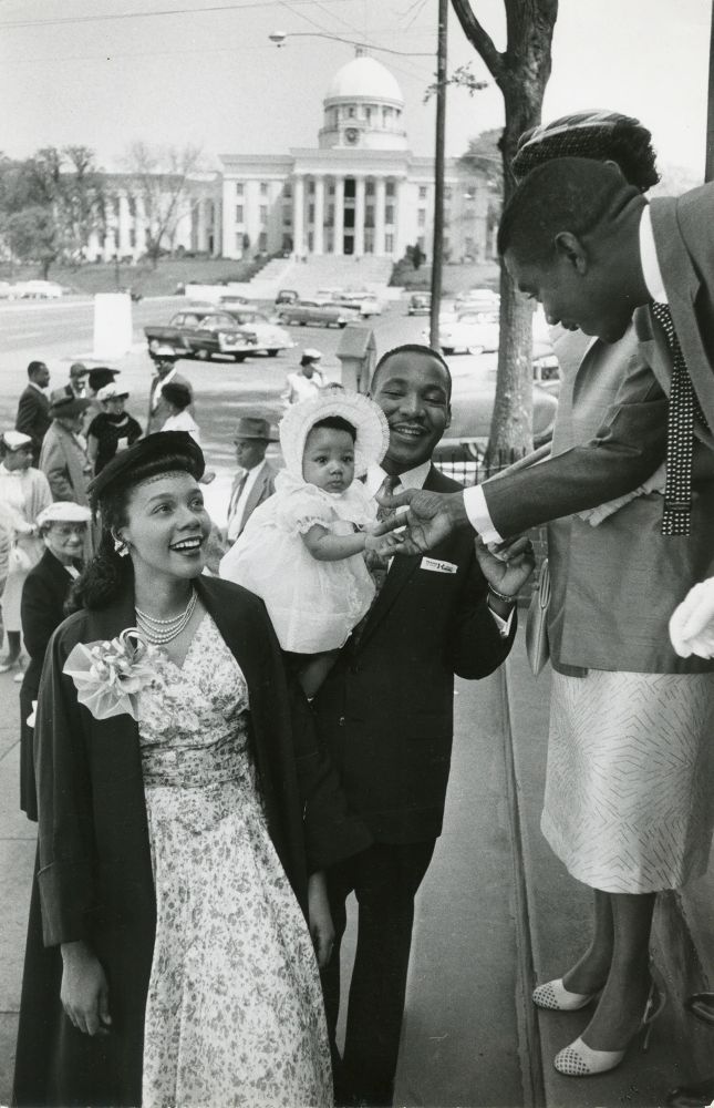 A fabulously dressed Coretta Scott King smiles next to Martin Luther King, who smiles and hold their child who has their tiny hand shaken by a larger hand 