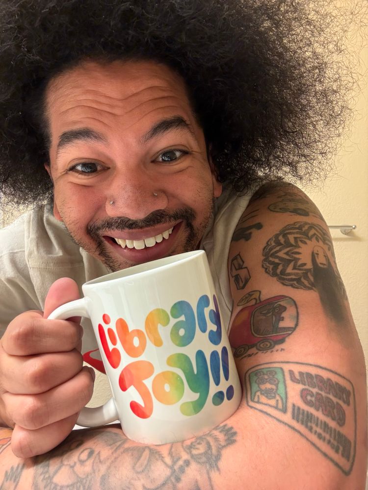 Mychal smiles with his tattoos showing and holds a Library Joy coffee mug