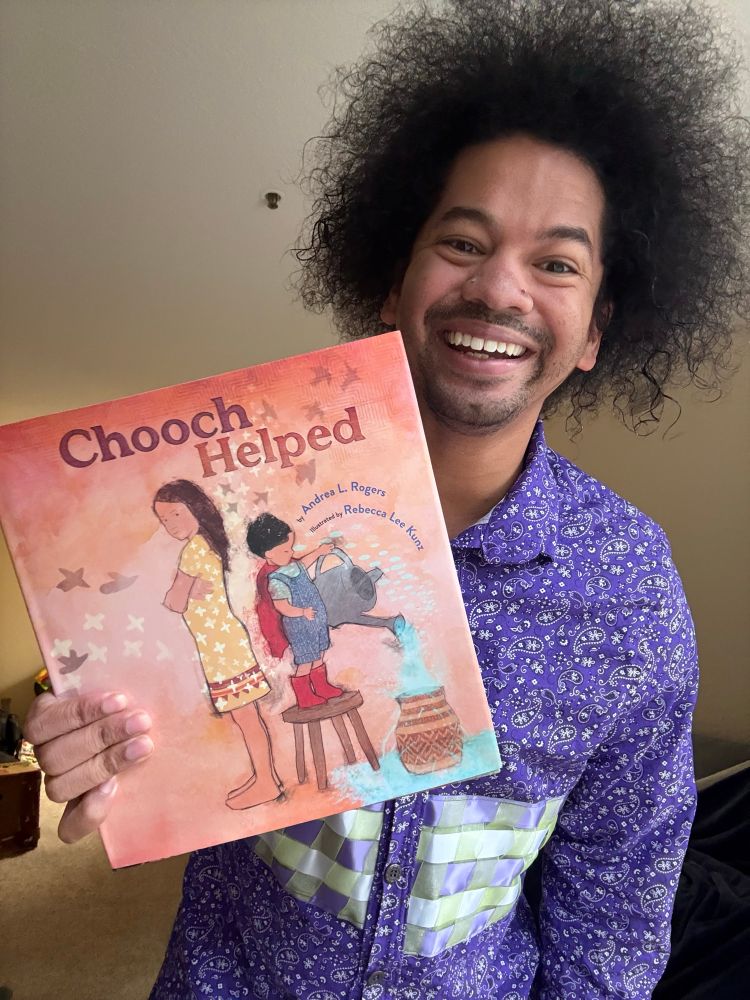 Mychal smiles in Indigenous purple shirt, holding the book CHOOCH HELPED by Andrea L. Rogers and illustrated by Rebecca Lee Kunz