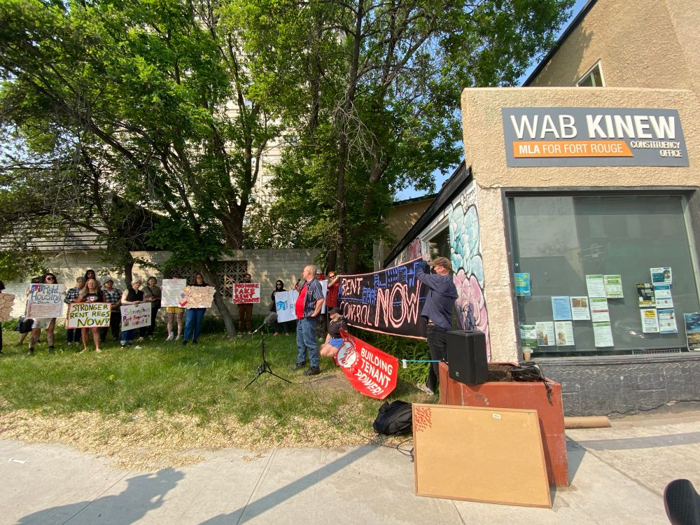 Protestors hold banner reading “rent control now” next to Wab Kinew’s office