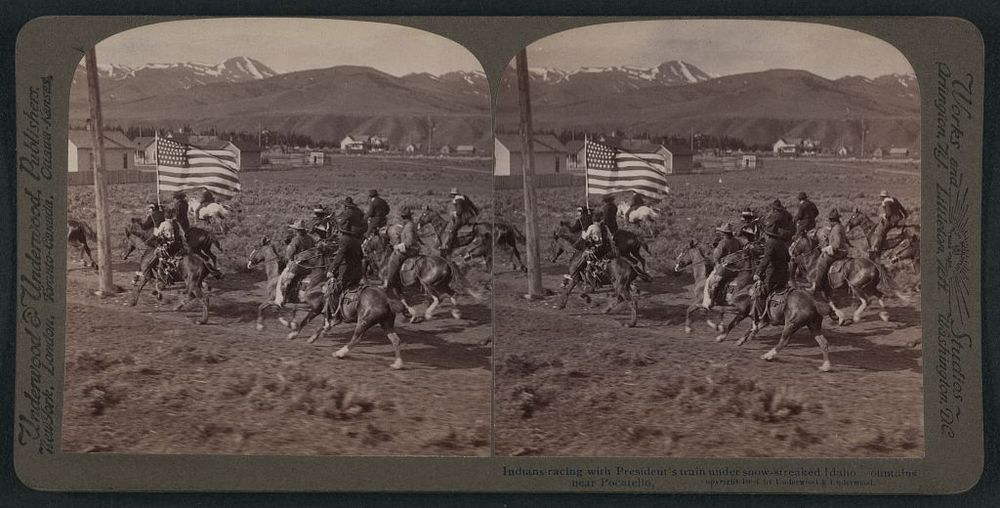 stereograph photo depicts Native Americans on horseback bearing an American flag, point of view as though the photographer is aboard a train coming up alongside them 
