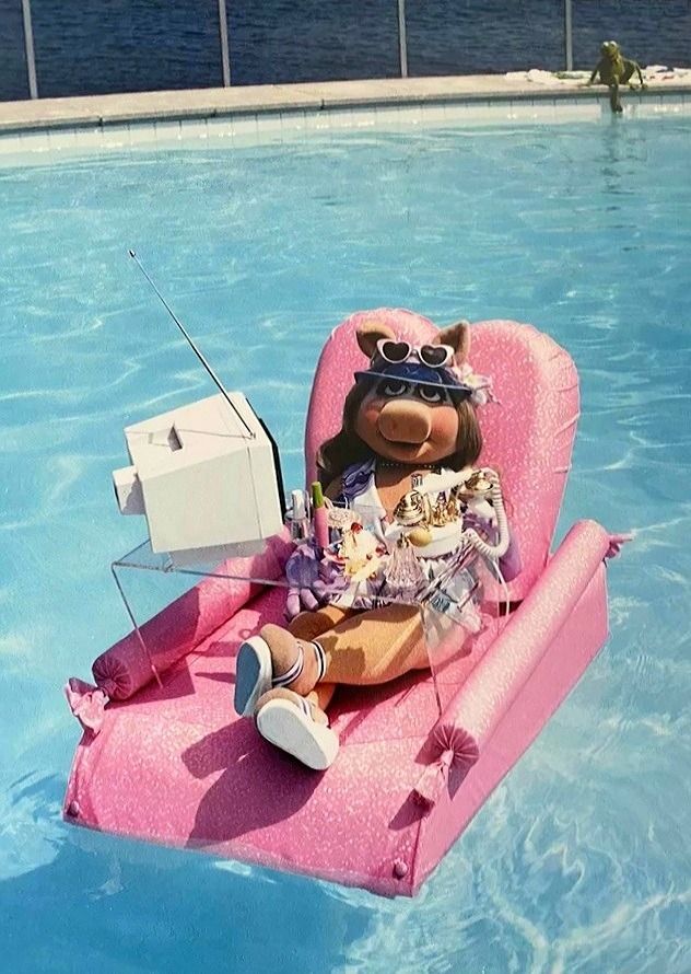Miss Piggy on a pink pool float with a heart shaped back, with a lap desk and portable television 