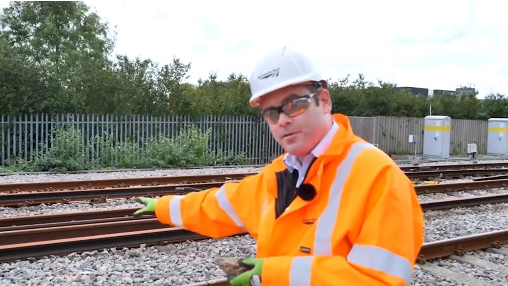 Photo of a main in a bright orange jacket and helmet standing in front of a railway line, pointing at the line with one hand and holding a lump of clay in the other 