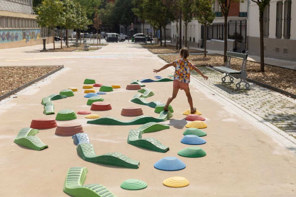 Ein Kind balanciert barfuß über bunte, modulare Balancier-Elemente, die wellenförmig und kreisförmig angeordnet sind. Der Parcours befindet sich auf einer autofreien, großzügigen Fläche in einem städtischen Umfeld. Es handelt sich vermutlich um eine temporäre oder dauerhaft umgestaltete Schulstraße. Das Bild zeigt eine bewegungsfreundliche, sichere und kreative Umgebung, in der Kinder frei spielen und sich entfalten können.