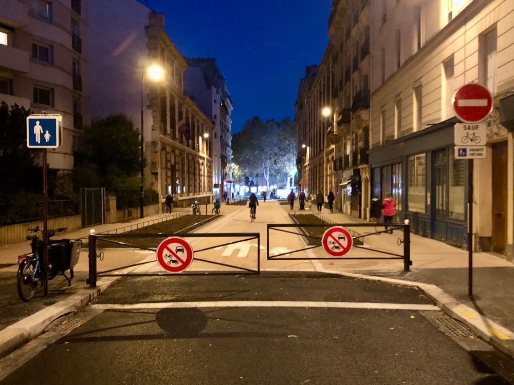 Foto einer neuen Schulstraßen in Paris, die nicht mehr für Autos befahrbar ist. Ein Schrankensystem versperrt die Zufahrt für die Autos. Menschen gehen auf dem Fußweg bzw. der Straße